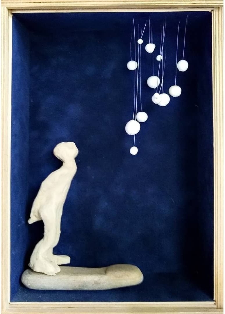 Small clay figure in a deep blue shadow box looking up at hanging white beads