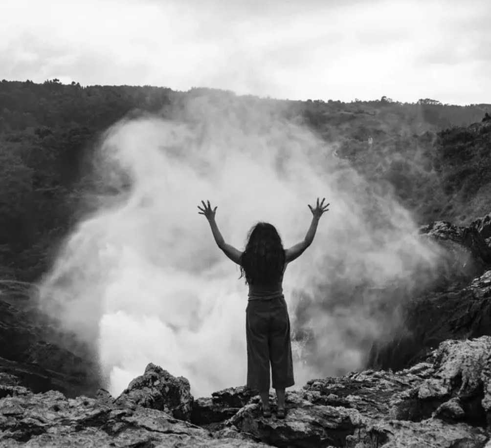 Hélène Desplechin standing on rocks with arms raised before a misting waterfall