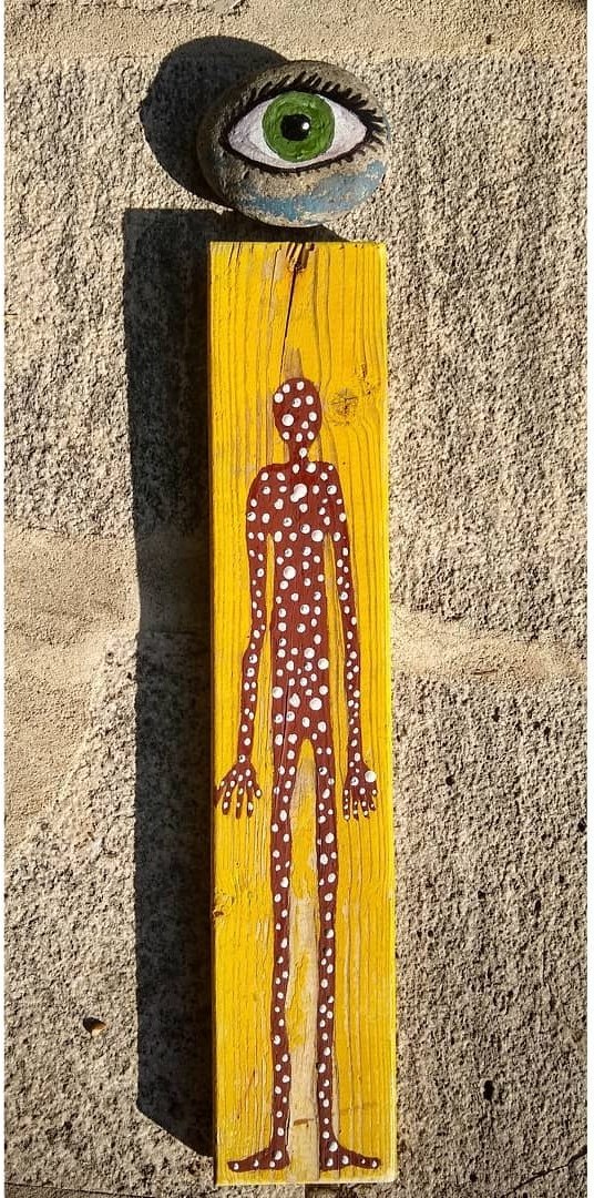 Painting of a polka-dotted figure on a yellow wooden board with a painted eye stone above